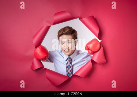 Ritratto di giovane imprenditore sorridente indossando guanti da boxe nel foro strappato su carta rossa Foto Stock