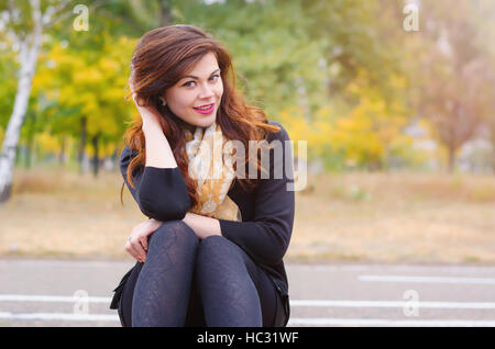 Ritratto di una giovane ragazza sorridente in una sciarpa che si siede su una panchina nel parco d'autunno. Autunno Caldo meteo e il sun. Foto Stock