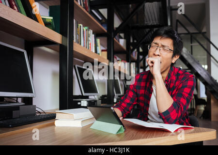 Foto di sbadigli asiatici studente maschio vestito in camicia in una gabbia e indossando occhiali che usano tablet presso la libreria. Foto Stock