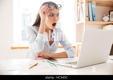 Foto di giovane imprenditrice in maglia bianca che indossano occhiali scioccante quando aprire un laptop. Foto Stock