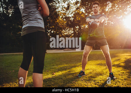 Giovane uomo facendo peso kettlebell allenamento con personal trainer femmina nel parco. Montare l'uomo bollitore oscillante campana con istruttore di fitness. Foto Stock