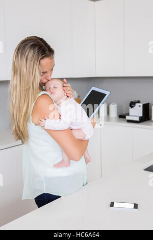 Madre utilizzando tavoletta digitale mentre si sta portando il suo bambino in cucina Foto Stock