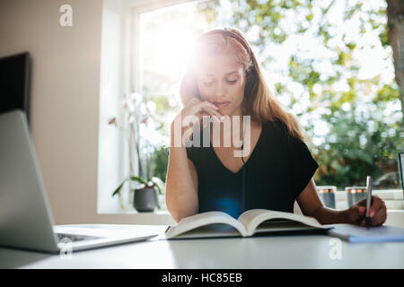 Giovane donna seduta a tavola con laptop e prendendo appunti nel libro. Studentessa studiare a casa. Foto Stock