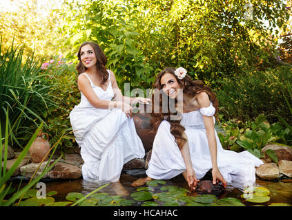 Due graziosi sorelle in vestiti bianchi presso il laghetto di ninfee. Le donne attingere acqua nella piccola brocca di argilla. Foto Stock