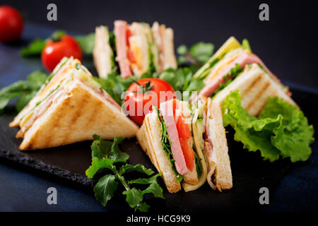 Club sandwich al prosciutto, pancetta, pomodori, cetrioli, formaggio, uova e erbe su sfondo scuro Foto Stock