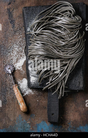 Materie non cotti nero inchiostro seppia spaghetti la pasta con la farina e il disco di pasta cutter su nero tagliere su legno scuro dello sfondo. Vista superiore con Foto Stock