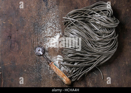 Materie non cotti nero inchiostro seppia spaghetti la pasta con la farina e il disco di pasta cutter su legno scuro dello sfondo. Vista da sopra con spazio per il testo. Foto Stock