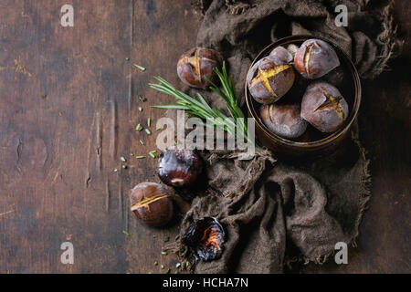 Castagne arrostite nella cenere con rosmarino in barattolo di latta su un letto di sacco su un vecchio legno scuro dello sfondo. Vista da sopra con spazio per il testo. Foto Stock