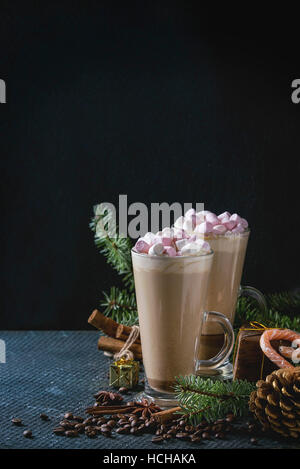 Due bicchieri di caffelatte con marshmallow rosa con decorazioni di Natale, caramelle, spezie, i chicchi di caffè e abete su texture scuro dello sfondo. Top vie Foto Stock