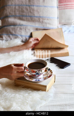Donna seduta sul letto,sfogliando un libro e bere una tazza di caffè. Foto Stock