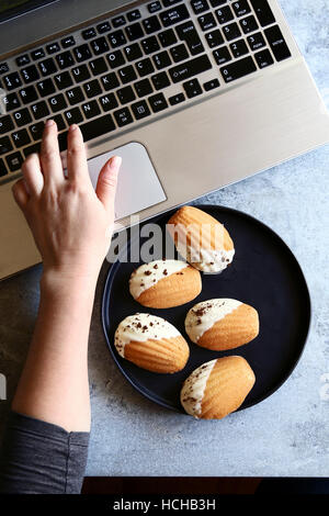 Donna seduta al tavolo,lavorando sul computer portatile e mangiare madeleines cookies.vista superiore Foto Stock