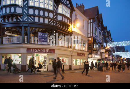 Tramonto nel centro città di Chester, High Cross, Cheshire, Regno Unito. Gli acquisti natalizi in pieno svolgimento nel costoso settore storico della vendita al dettaglio del centro città Tudor. Architettura Tudor precoce nelle Chester Rows che consistono in passaggi coperti al primo piano dietro i quali si trovano gli ingressi di negozi e altri locali, Foto Stock