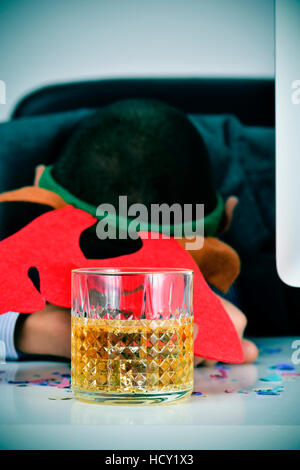 Primo piano di un giovane imprenditore caucasico indossando una renna archetto di corna a dormire la sua scrivania, coperto di coriandoli e con un bicchiere di liquore in Foto Stock