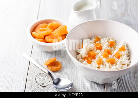 Prima colazione da cottage cheese con fettine di albicocche secche in una ciotola bianco su bianco sullo sfondo di legno Foto Stock