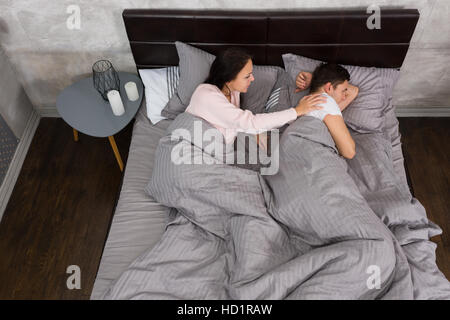 Giovane donna wake up offeso il suo marito mentre giaceva nel letto e indossando pigiami, vicino comodino con candele in camera da letto in stile loft con Foto Stock