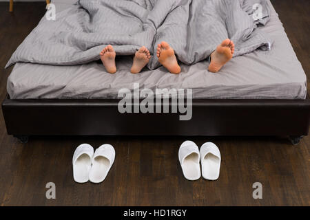 Close up di maschio e femmina piedi sotto coperta grigio in stile moderno bed in prossimità di due paia di pantofole bianco Foto Stock