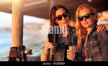 Ritratto di due belle ragazze trascorrere il tempo libero in outdoor cafe con bevande, modelli indossando occhiali da sole elegante, godendo luminosa giornata di sole Foto Stock