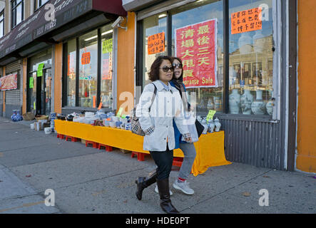 Due donne asiatiche camminando a braccetto sulla strada principale e passando un negozio di sconti in Chinatown, lavaggio. Queens, a New York City. Foto Stock