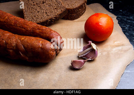 Vista ravvicinata salsiccia affumicata con segale pane nero, molti pomodori e aglio per il marrone del confezionamento della carta. Foto Stock