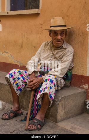 93 anni di vecchio uomo Maya in abito tradizionale si trova sulla strada di San Pedro La Laguna, Guatemala. Seduti sul cordolo con la sua canna da zucchero nella parte anteriore di un pastello pa Foto Stock