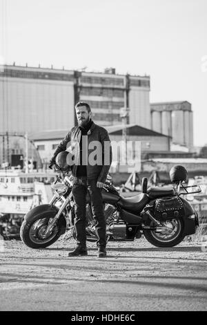 A piena lunghezza Ritratto di biker holding casco mentre in piedi da moto contro l'impostazione industriale Foto Stock