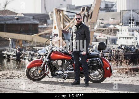 A piena lunghezza Ritratto di biker in piedi da moto contro l'impostazione industriale Foto Stock
