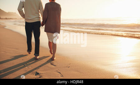 Vista posteriore mozzato colpo di una coppia senior tenendo le mani di camminare sulla spiaggia. Coppia matura insieme facendo una passeggiata sulla riva del mare al tramonto. Foto Stock