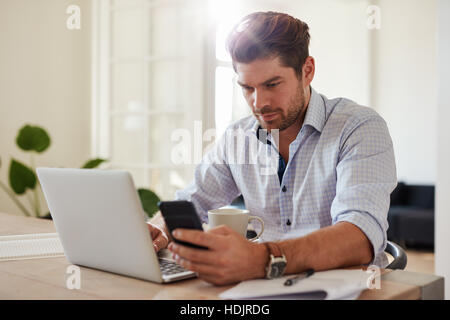 Colpo di giovane uomo che lavora con il computer portatile e il telefono cellulare al suo ufficio di casa. Bel maschio seduta a tavola tenendo lo smartphone e utilizzando il computer portatile Foto Stock