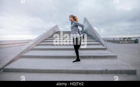 Vista laterale della giovane donna che si allunga la gamba all'aperto. donna in forma fisica che si prepara a salire le scale durante l'allenamento. Foto Stock
