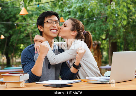 Carino giovane donna seduta e baciare allegro asian giovane uomo in vetri esterni Foto Stock