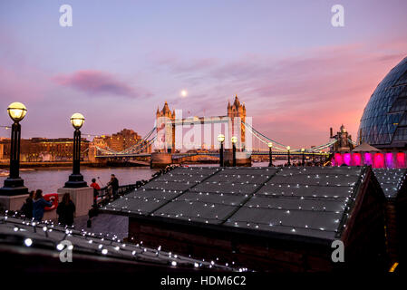 London Tower Bridge in una splendida luna piena serata con il mercatino di Natale di capanne con luci in primo piano. Foto Stock