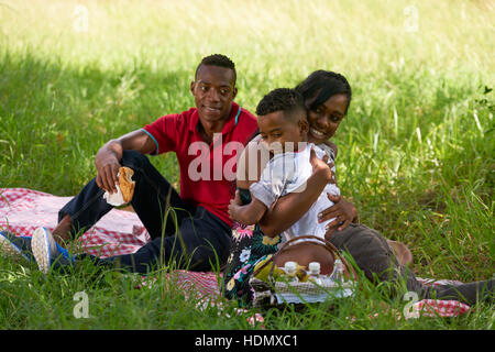 Felice coppia nero con figlio nel parco della città. Famiglia americana africana con un giovane uomo, donna e bambino facendo picnic, avendo divertimento all'aperto. Foto Stock