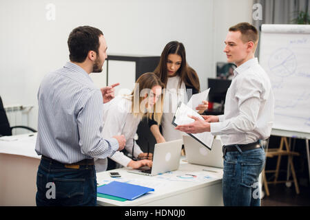 Quattro giovani uomini di affari che lavorano come un team riuniti intorno al computer portatile in un piano aperto ufficio moderno Foto Stock