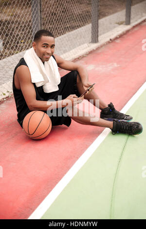 Foto di giovani africani allegro giocatore di basket seduta nel parco vicino la pallacanestro e tenendo compressa. Guarda la fotocamera. Foto Stock