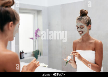 Felice giovane donna di applicare la maschera per il viso in bagno e sorridente. Bellissima femmina nella parte anteriore dello specchio facendo un trattamento di bellezza. Foto Stock