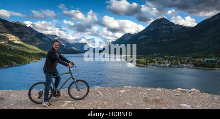 Un uomo si siede su una bicicletta a bordo dell'acqua con le montagne rocciose e un lago in background, Parco Nazionale dei laghi di Waterton; Alberta, Canada Foto Stock
