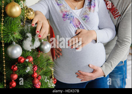 Incinta giovane nei pressi di un albero di Natale. Il marito abbracciando la sua moglie incinta. Ragazza decora un albero di Natale. Foto Stock