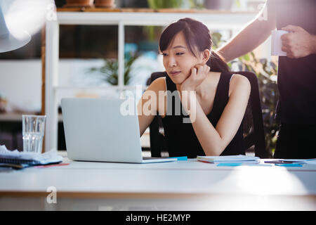 Colpo di asian giovane donna seduta alla sua scrivania che lavora sul computer portatile con la collega di sesso maschile in attesa. Foto Stock