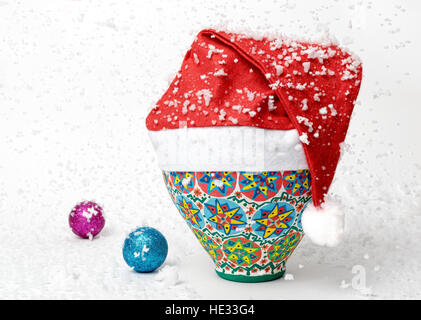 Egiziano colorato decorato vaso in ceramica indossa Santa Claus Red Hat su sfondo bianco con due colori splendenti palle di Natale e la caduta di neve Foto Stock