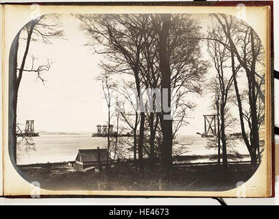 Questa fotografia storica mostra la costruzione del Forth Bridge da Port Edgar, catturando la monumentale impresa ingegneristica. L'immagine evidenzia gli alberi circostanti, aggiungendo un contesto al paesaggio dell'epoca. La foto riflette sia il progresso industriale che l'ambiente naturale del tempo. Foto Stock