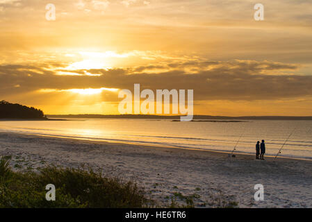 Due uomini stand la pesca e la visione di un luminoso giallo ed arancione tramonto su Callala Bay sul Nuovo Galles del Sud costa sud dell'Australia Foto Stock