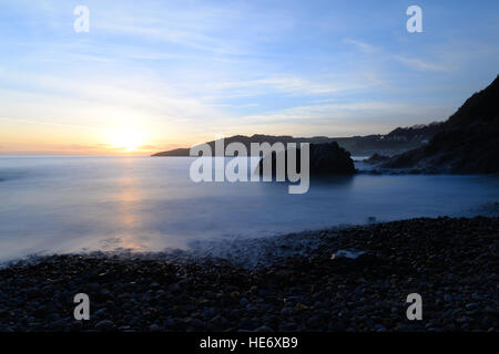 Tramonto con l'alta marea in una baia riparata sulla penisola di Gower. Una grande roccia sembra appesa al mare resa nebbiosa dall'esposizione temporizzata Foto Stock