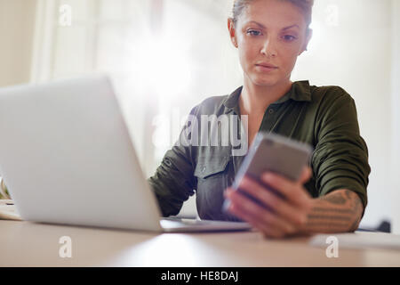 Colpo di attraente giovane donna seduta a tavola con il computer portatile e la lettura di un messaggio di testo sul suo telefono cellulare Foto Stock
