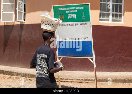 Un giovane uomo porta un cesto della biancheria sulla testa oltre un cartello per il "Rehabilitation and Community Based Poverty Reduction Project" (RCPRP), Ufficio del Ward 144, a Kabala, Distretto di Koinadugu, Sierra Leone. Foto Stock
