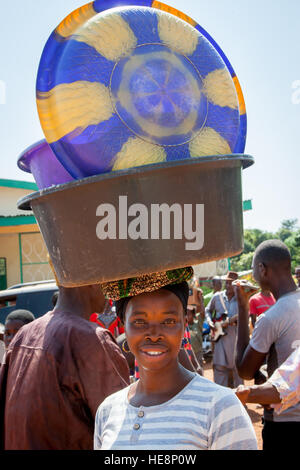 Una donna sorridente bilancia un grande contenitore di plastica a motivi blu e gialli sulla testa, passando davanti a una performance musicale a Kabala, Sierra Leone. Foto Stock