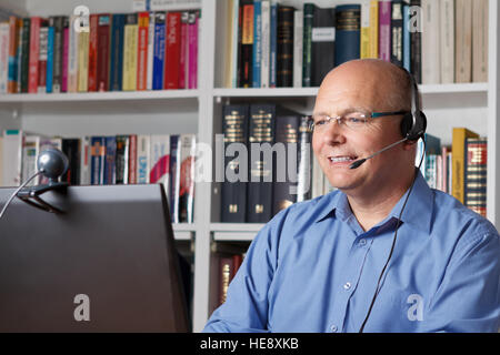 Senior sorridente Uomo con cuffie, computer e webcam a parlare con la sua famiglia via internet, libri sugli scaffali in background Foto Stock