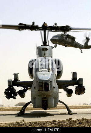 Un US Marine Corps (USMC) AH-1W Super Cobra elicottero armato di AGM-114 Hellfire missili e 2,75 pollici aletta pieghevole antenna (Razzi FFAR) attende il rifornimento di carburante in corrispondenza di una zona di avanzamento Punto di rifornimento (FARP), a Tallil Air Base, Iraq durante l'Operazione Iraqi Freedom. Un esercito americano (USA) UH-60 Black Hawk elicottero è in avvicinamento in background. Foto Stock