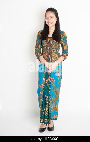 Ritratto di giovane sud-est ragazza asiatica tradizionale Malay kebaya batik abito sorridente a piena lunghezza permanente sulla pianura dello sfondo. Foto Stock