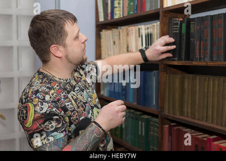 Un uomo con una barba sceglie un libro in biblioteca. Foto Stock