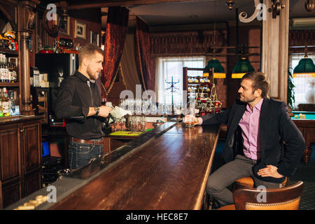 Sorridente attraente giovane barista tergi bicchieri e a parlare con l'uomo in bar Foto Stock